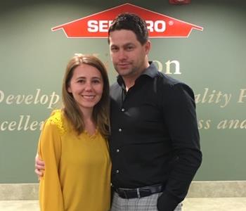 Woman with brown hair and mustard shirt with a man with dark hair and a black shirt standing in front of a SERVPRO sign.